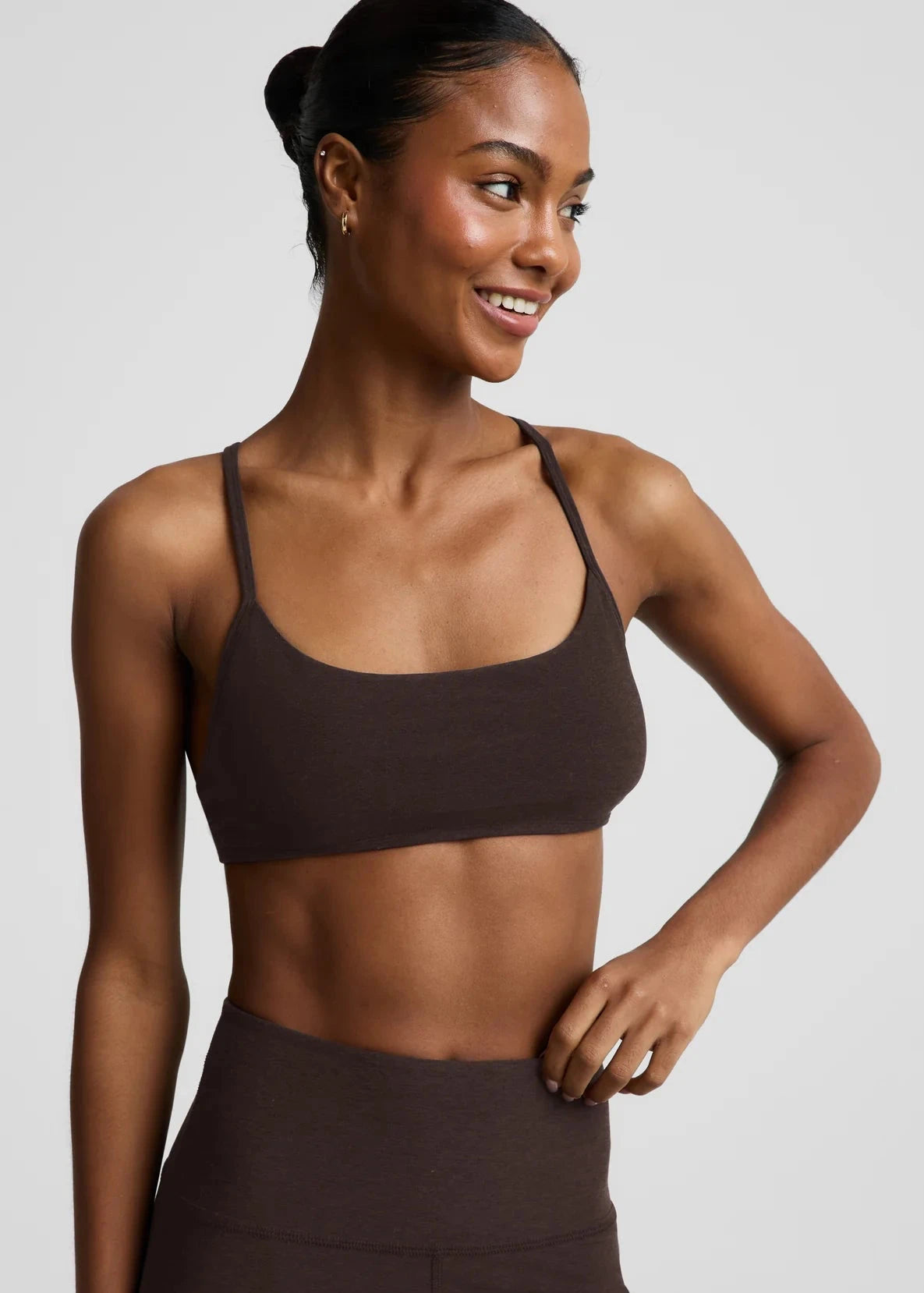 A woman wearing a dark brown scoop-neck sports bra and matching high-waisted leggings, photographed against a solid light gray studio background.