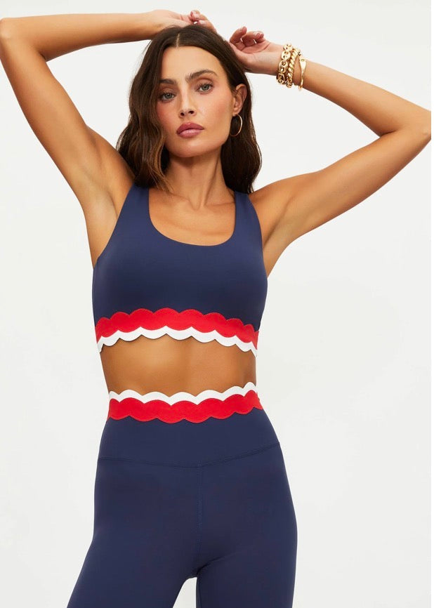 Woman wearing a navy sports bra with red and white accents and matching leggings on a light gray background.