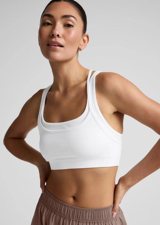 Woman wearing a white sports bra and brown pants on a light gray background